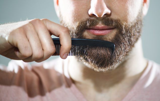 Closeup of unrecognizable caucasian man combing his beard and mustach with small black comb.He has neat,fully grown brown beard and mustache.Studio shot over gray background. ,=Front view.