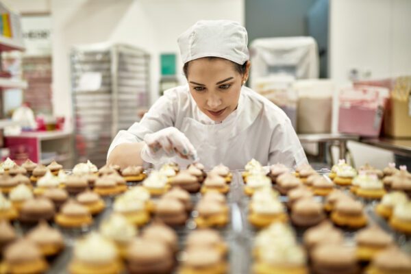 Low angle view of focused baker in mid 30s decorating fresh batch of vegan cupcakes in commercial kitchen.