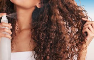 women with long curly brown hair spraying a mist into her hair in front of blue background