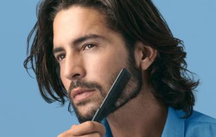 young white man with long brown hair combing his beard with blue background