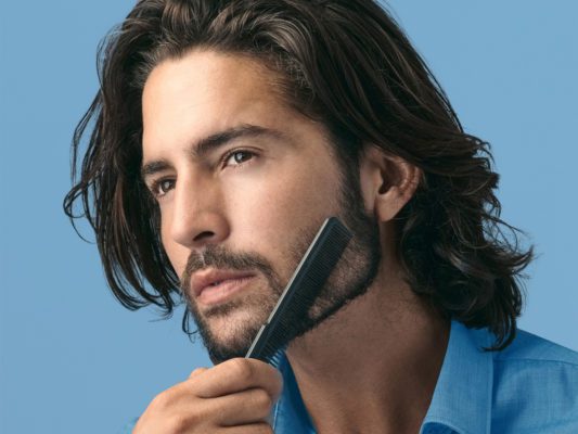 young white man with long brown hair combing his beard with blue background